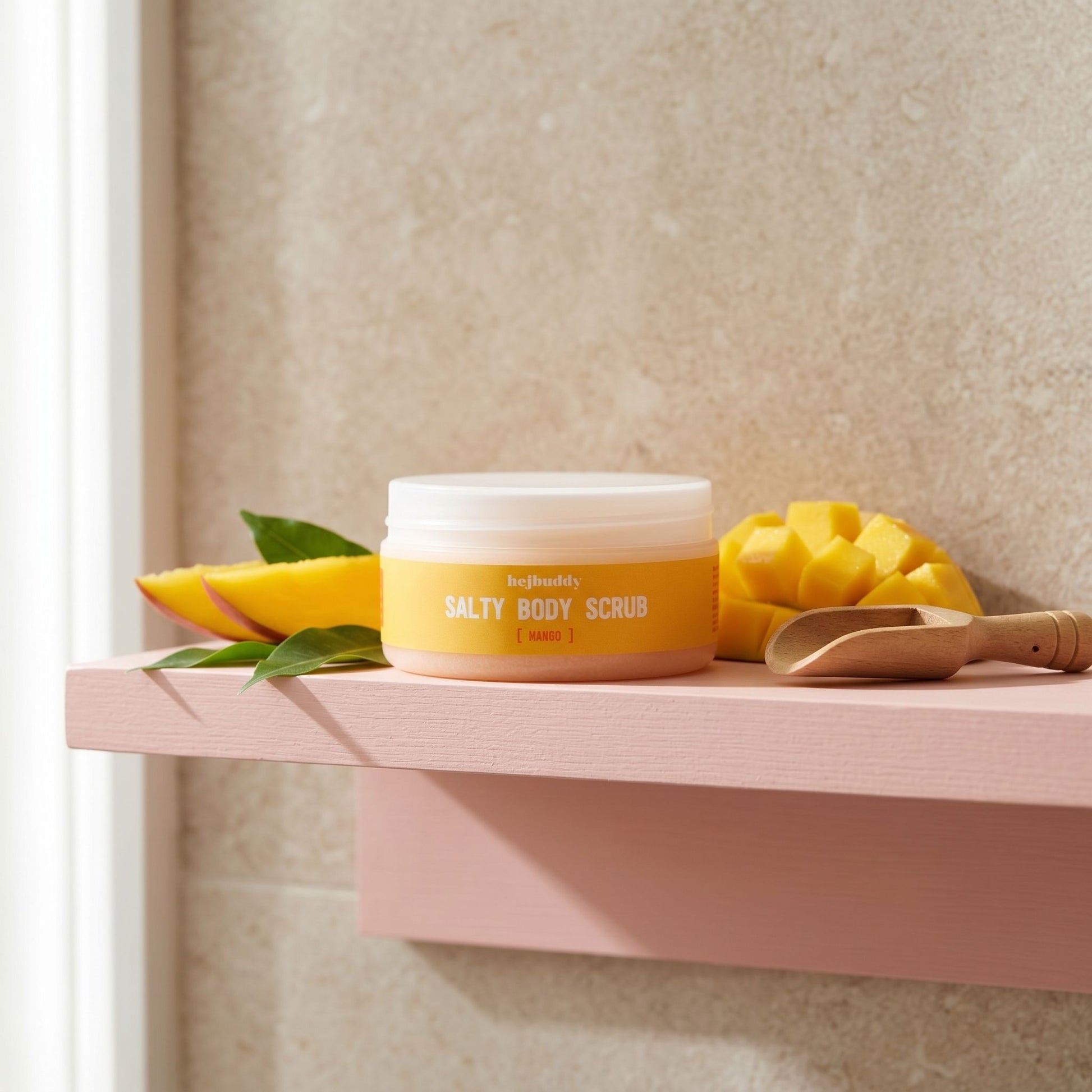 HejBuddy Mango Salty Body Scrub container on a pink bathroom shelf with a stone wall background and natural lighting.