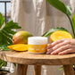 Lotion jar on a balcony with lush greenery and mangoes, sunlight highlighting the creamy texture of the bottle.